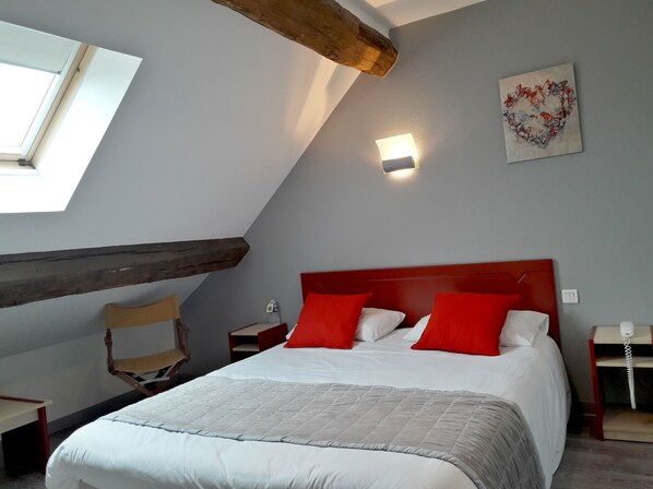 A bedroom with a sloped ceiling, a bed with white and red bedding, a wooden headboard, a nightstand with a lamp, and a wall-mounted light. - Hôtel Relais du Lac (Villegusien-le-Lac)