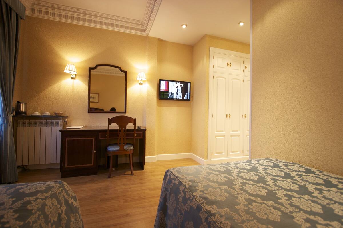 Una camera d'albergo con una scrivania in legno, una sedia, una televisione a parete e due letti con coperte decorate.