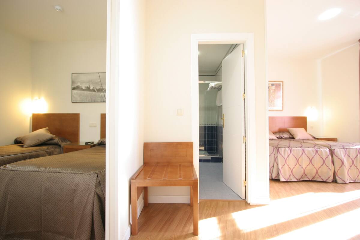 Una camera d'albergo con due letti, una panca di legno e un bagno visibile attraverso una porta aperta.