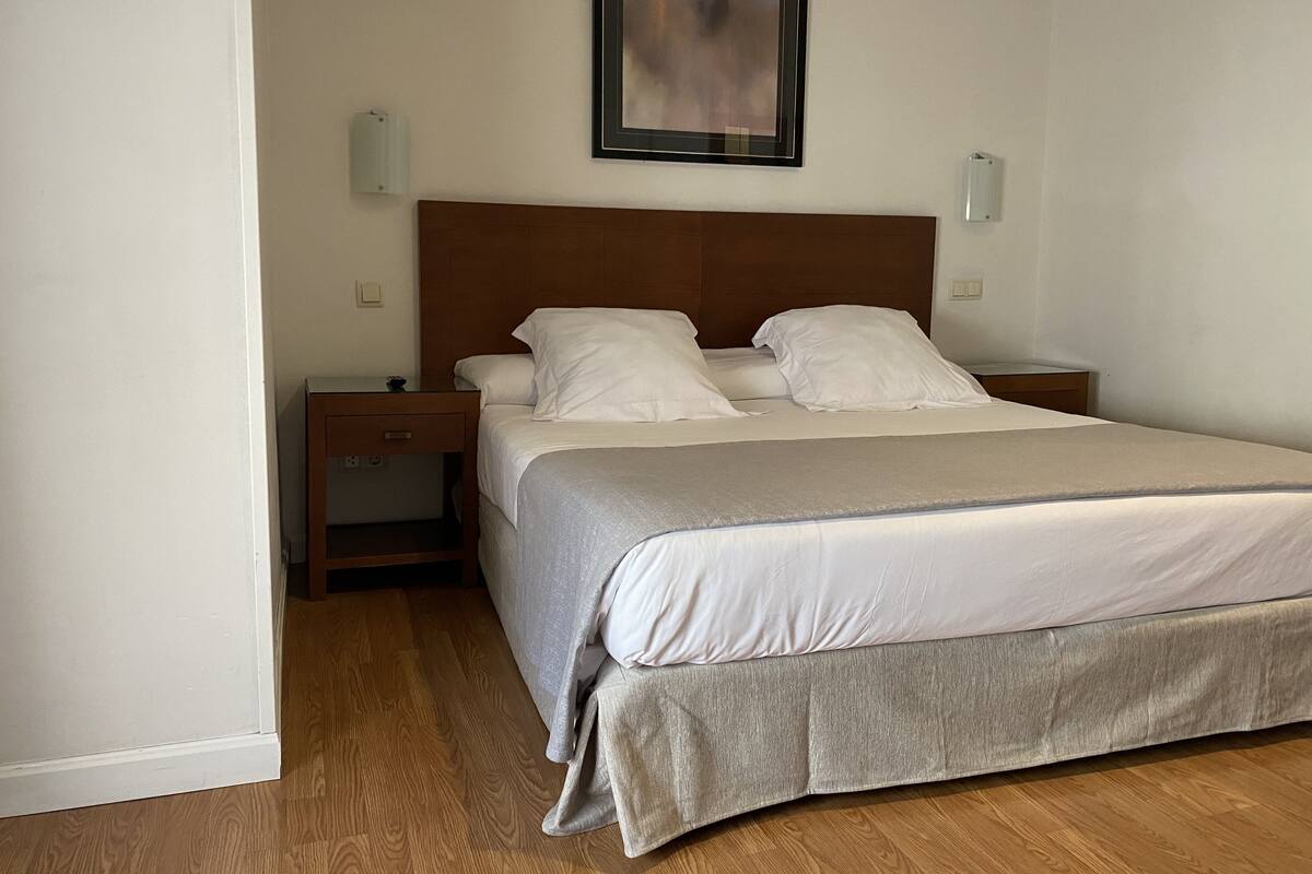 Un letto ben rifatto con una coperta grigia e cuscini bianchi, un comodino in legno con una lampada e un quadro appeso al muro.