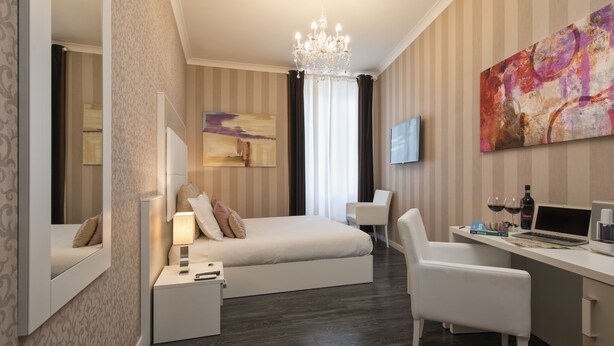 A modern hotel room with a bed, bedside table, lamp, desk with laptop, chair, TV, and artwork on the wall. - San Pietro Suites (Rome)