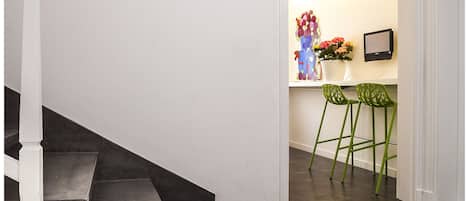 A modern interior with a staircase, a white table with magazines, and a kitchen area with stools and a vase of flowers.