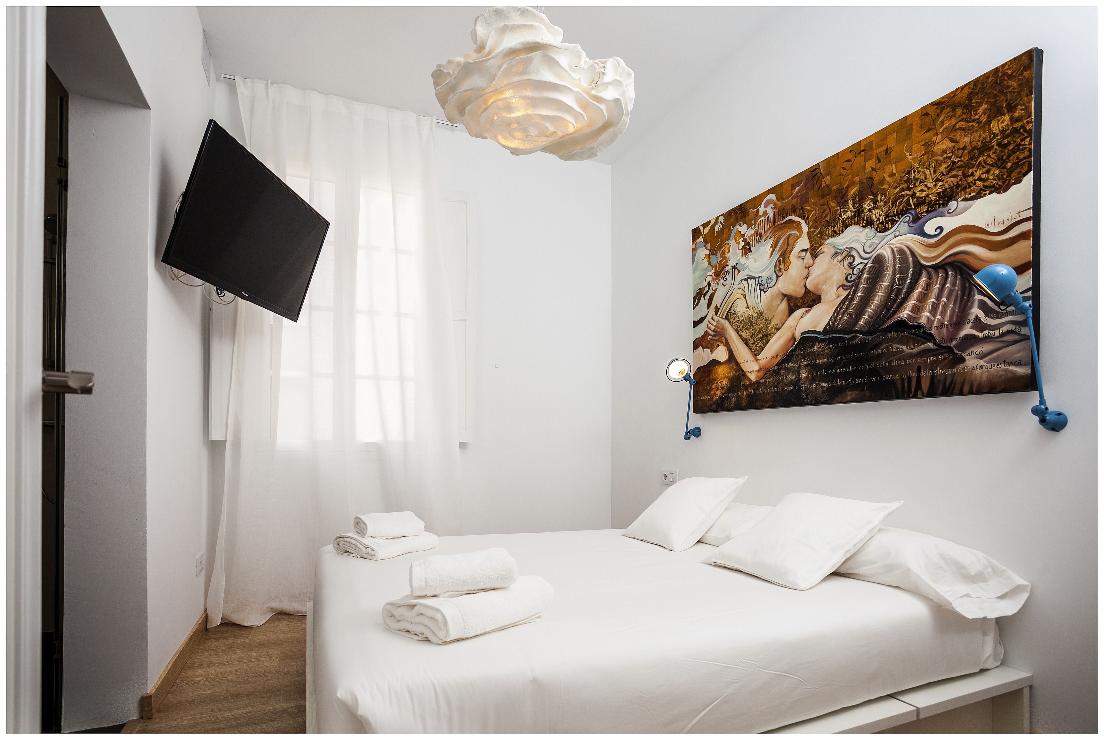 A hotel room with a bed, white curtains, a mounted TV, a painting on the wall, and a ceiling light fixture.