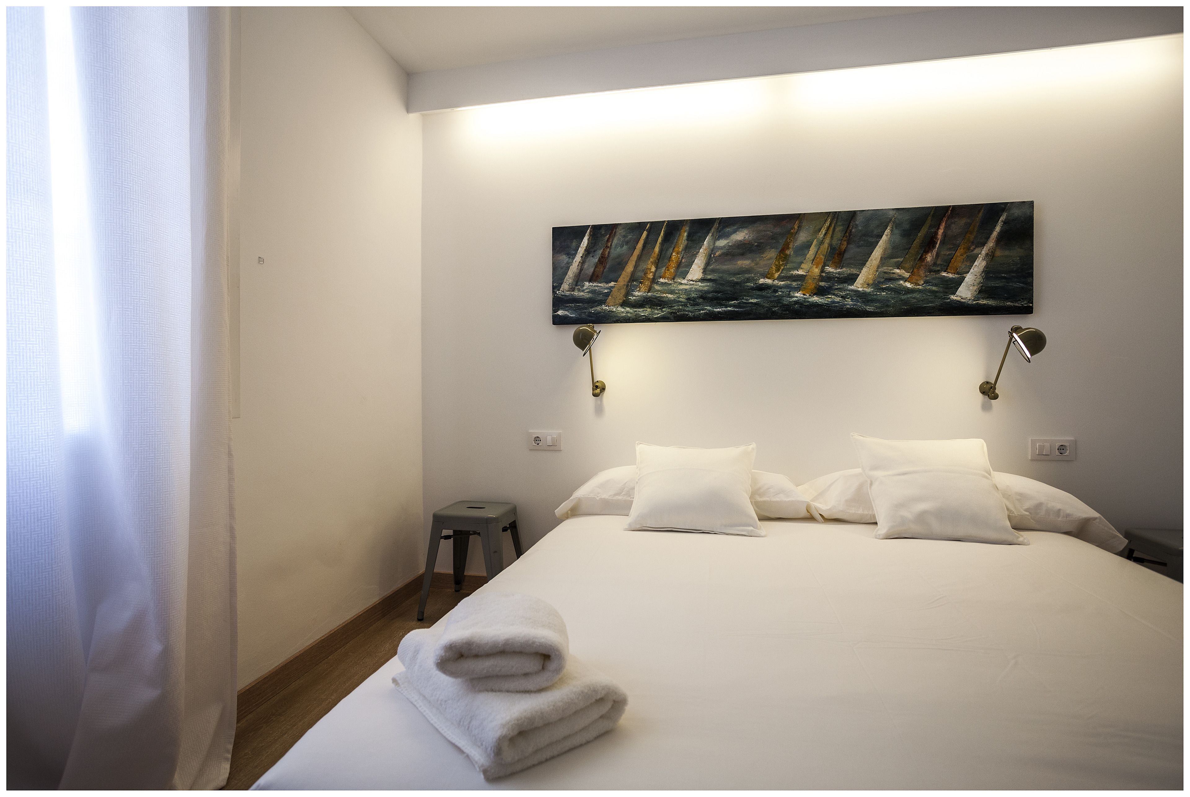 Una cama bien hecha con sábanas y almohadas blancas, un cuadro con barcos de vela en la pared y un taburete al lado de la cama.