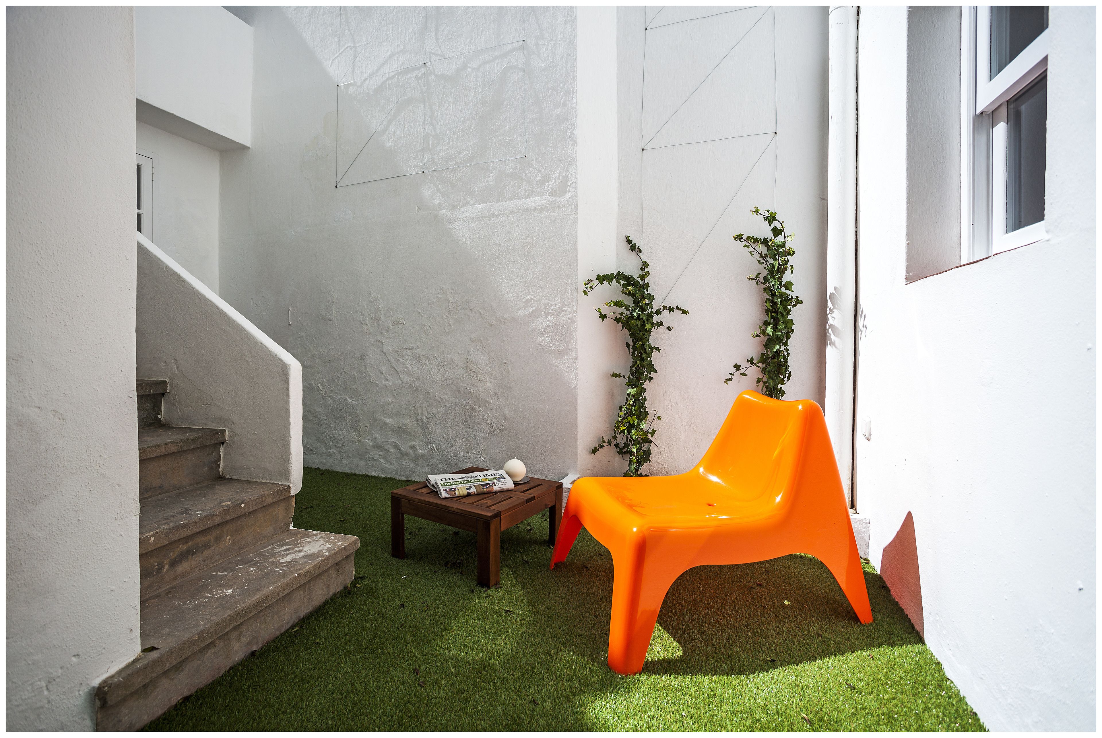Un pequeño espacio al aire libre con una escalera de madera, una mesa de madera con una revista, una silla de plástico naranja y una planta en maceta.