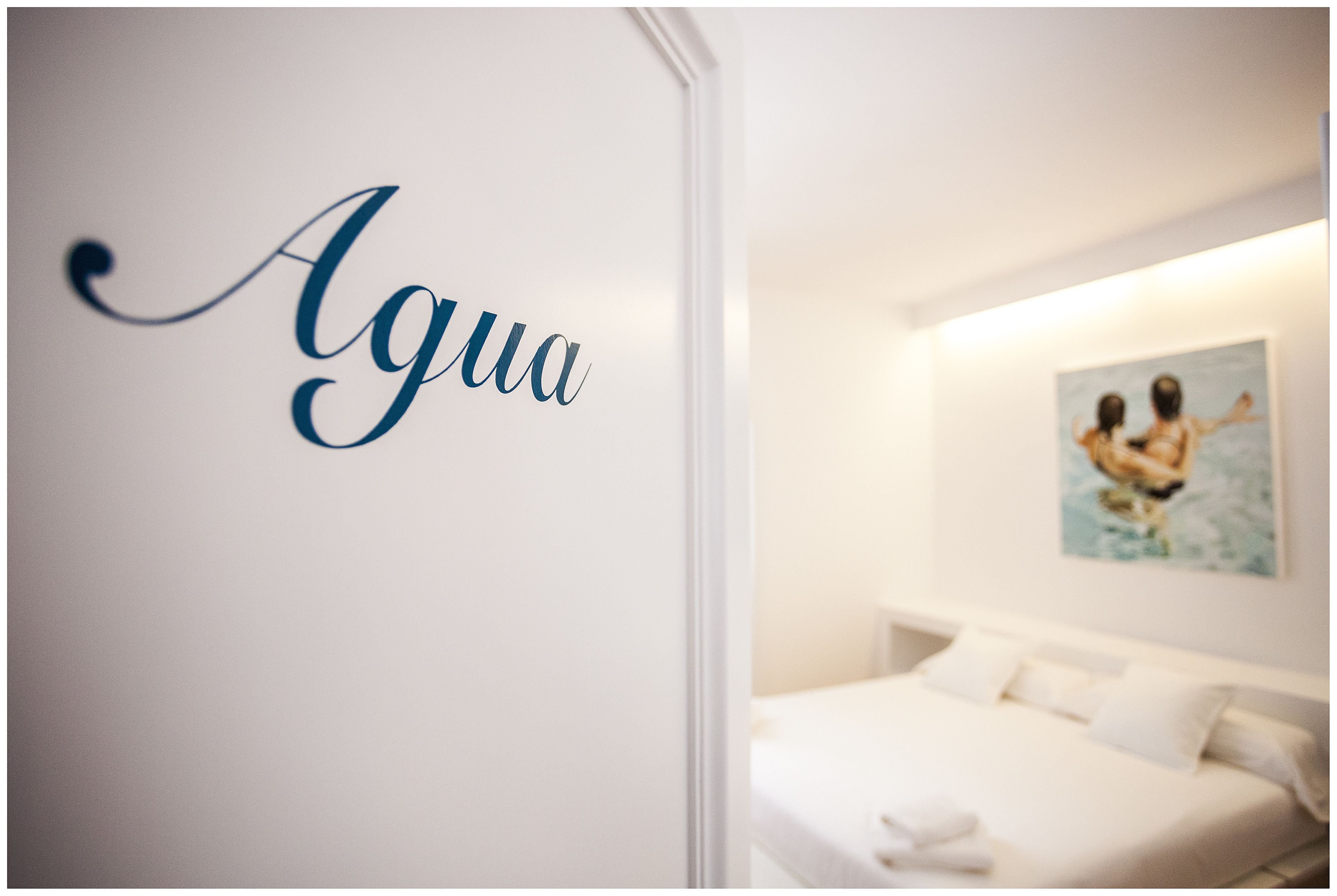 Una habitación con cama, pared con la palabra 'Agua' en cursiva y un cuadro de dos personas en el agua.