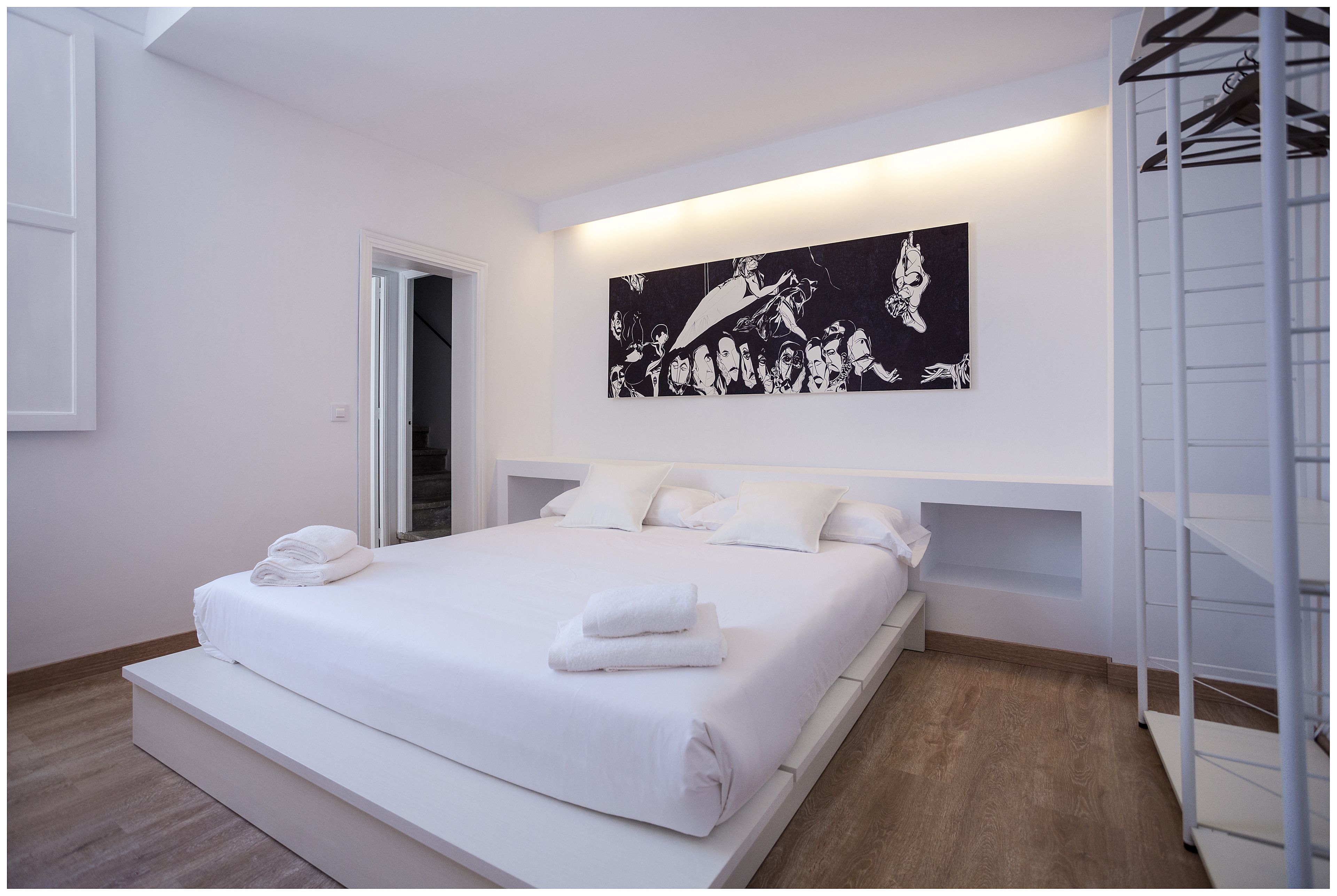 A modern bedroom with a large bed, white linens, a black and white artwork above the bed, and a white wardrobe with a mirror.