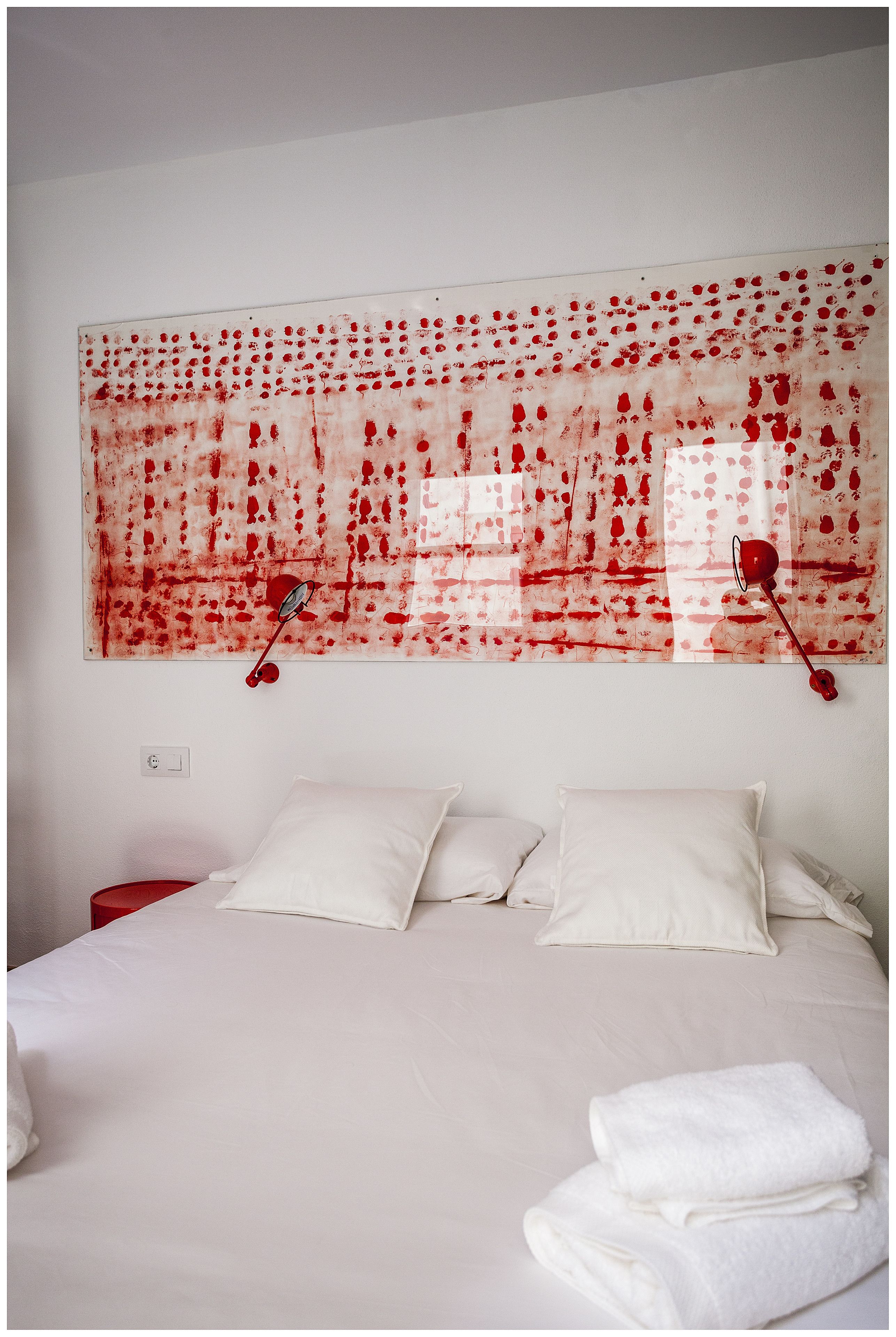 Habitación de hotel con un cuadro abstracto grande en tonos rojo y blanco, una cama con sábanas blancas y dos lámparas de noche rojas.