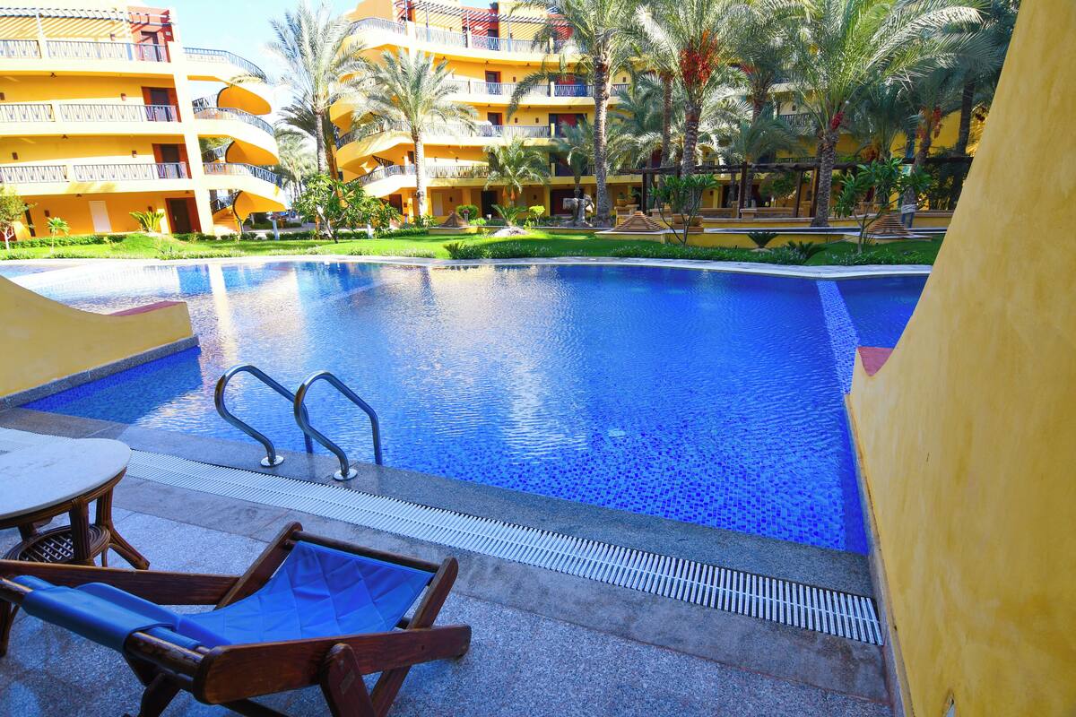 Piscina d'hotel con acqua cristallina, circondata da palme e un edificio giallo con balconi.