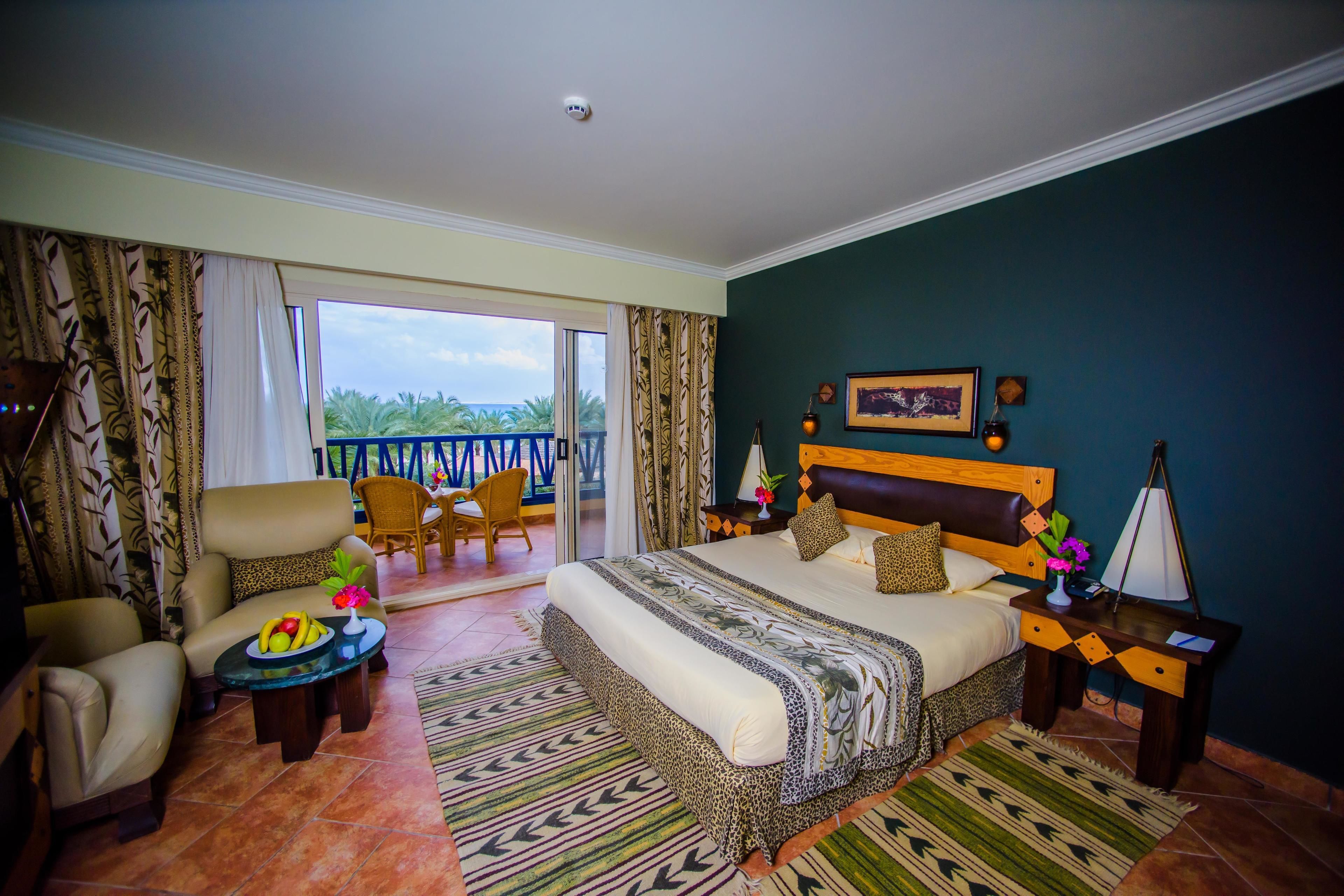 A hotel room with a large bed, a sofa, a dining table with fruit, and a balcony with chairs.