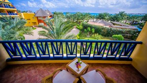 A balcony with wicker chairs and a small table, overlooking a resort area with palm trees and a pool. - Carols Beau Rivage Hotel (Marsa Matrouh)