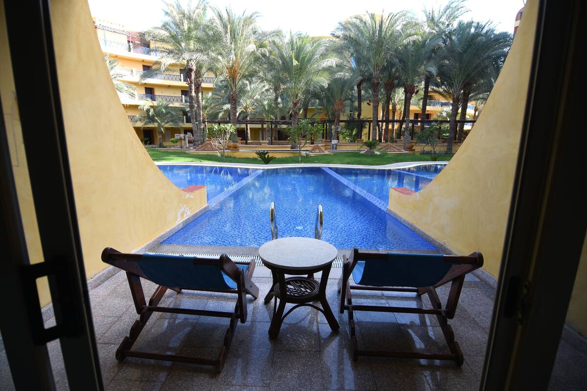 Vista di una piscina con due sedie e un tavolo in primo piano, circondata da palme e un edificio con balconi sullo sfondo.