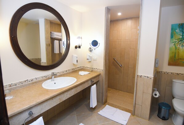 A bathroom with a sink, mirror, towel rack, shower, toilet, and a trash bin.
