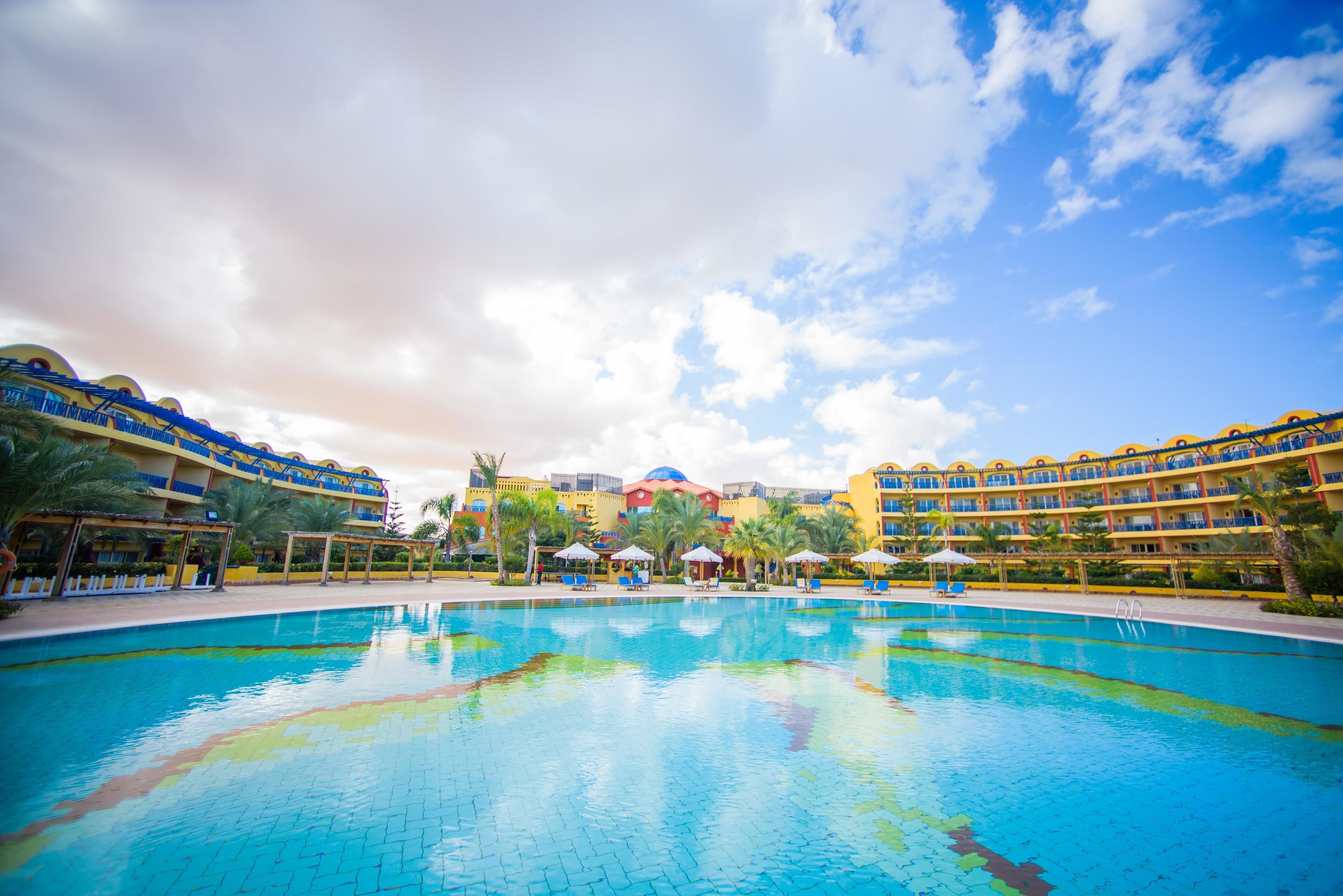 Zona de piscina de hotel con cielo despejado, varios niveles de balcones y un edificio central con cúpula.