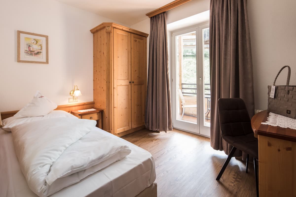 A bedroom with a bed, wooden wardrobe, desk, chair, and a view through a glass door.