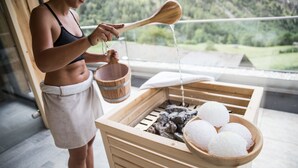 A person pouring water from a wooden ladle into a wooden bucket, with a view of a green landscape through a window. - Hotel Burgaunerhof (Martello)
