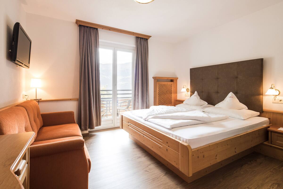 A hotel room with a bed, a sofa, a TV, and a balcony with curtains.
