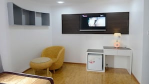 A room with a television mounted on the wall, a small round table, a wicker chair, and a desk with a lamp. - Hotel My World (Mandalay)