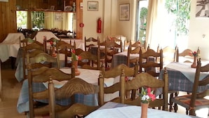 A hotel dining area with wooden chairs and tables, a vase with flowers on each table, and a view of the outdoors through large windows. - Karis Hotel (Kos)