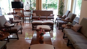 A spacious living room with a large TV on a stand, comfortable seating including sofas and armchairs, a coffee table with a vase of flowers, and large windows offering a view of greenery outside. - Karis Hotel (Kos)