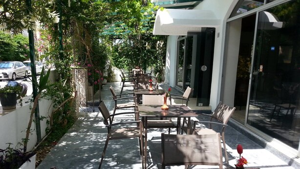 An outdoor seating area with tables and chairs, surrounded by greenery and a glass door entrance. - Karis Hotel (Kos)