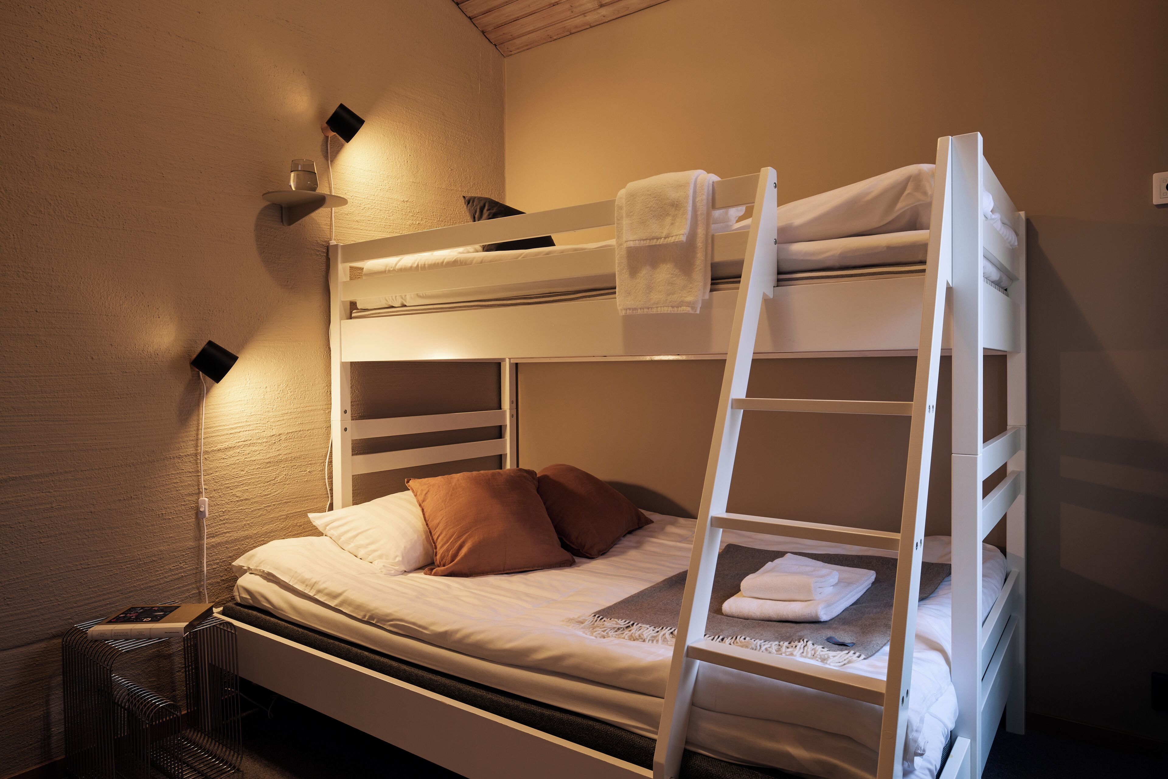 A bunk bed with a ladder, a bedside table with a lamp, and a wall-mounted shelf with a glass and a book.