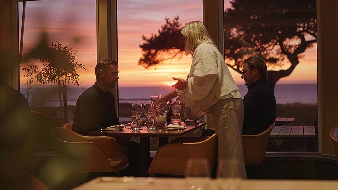 A dining area with a table set for four, large windows with a view of the sunset, and a potted plant on the windowsill.