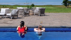 A swimming pool with two people sitting on the edge, lounge chairs, and a view of the sea. - Barsebäck Resort Hotell (Loddekopinge)