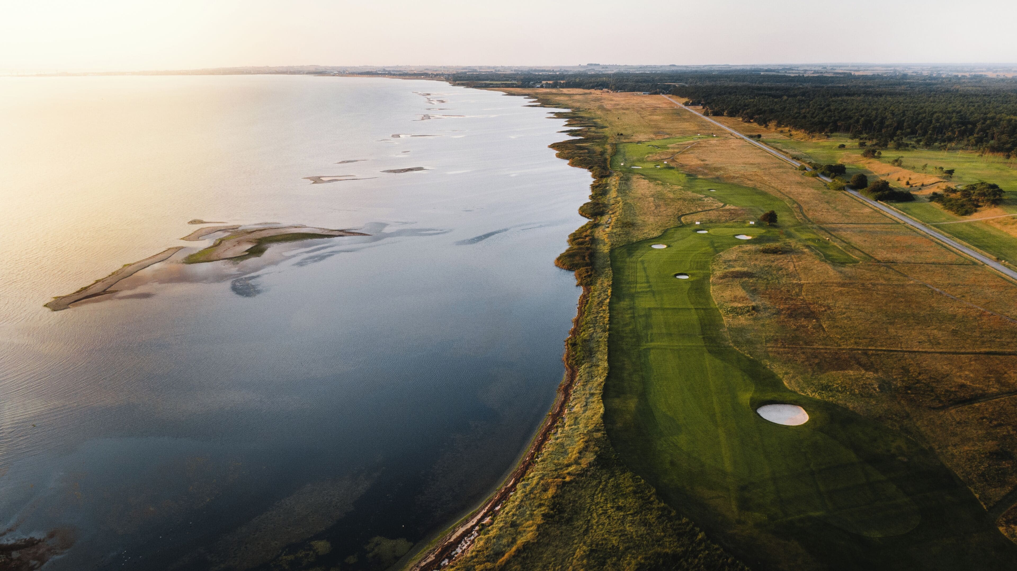 Vista aérea de un campo de golf costero con una masa de agua, una carretera y una zona boscosa.