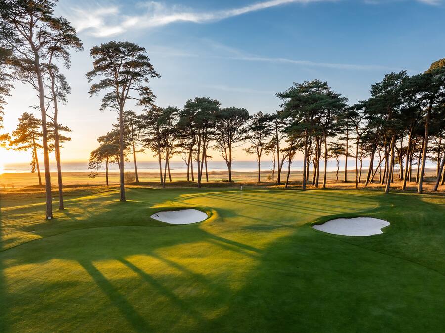 Un campo de golf con dos bunkers de arena, rodeado de árboles altos y un cielo despejado.
