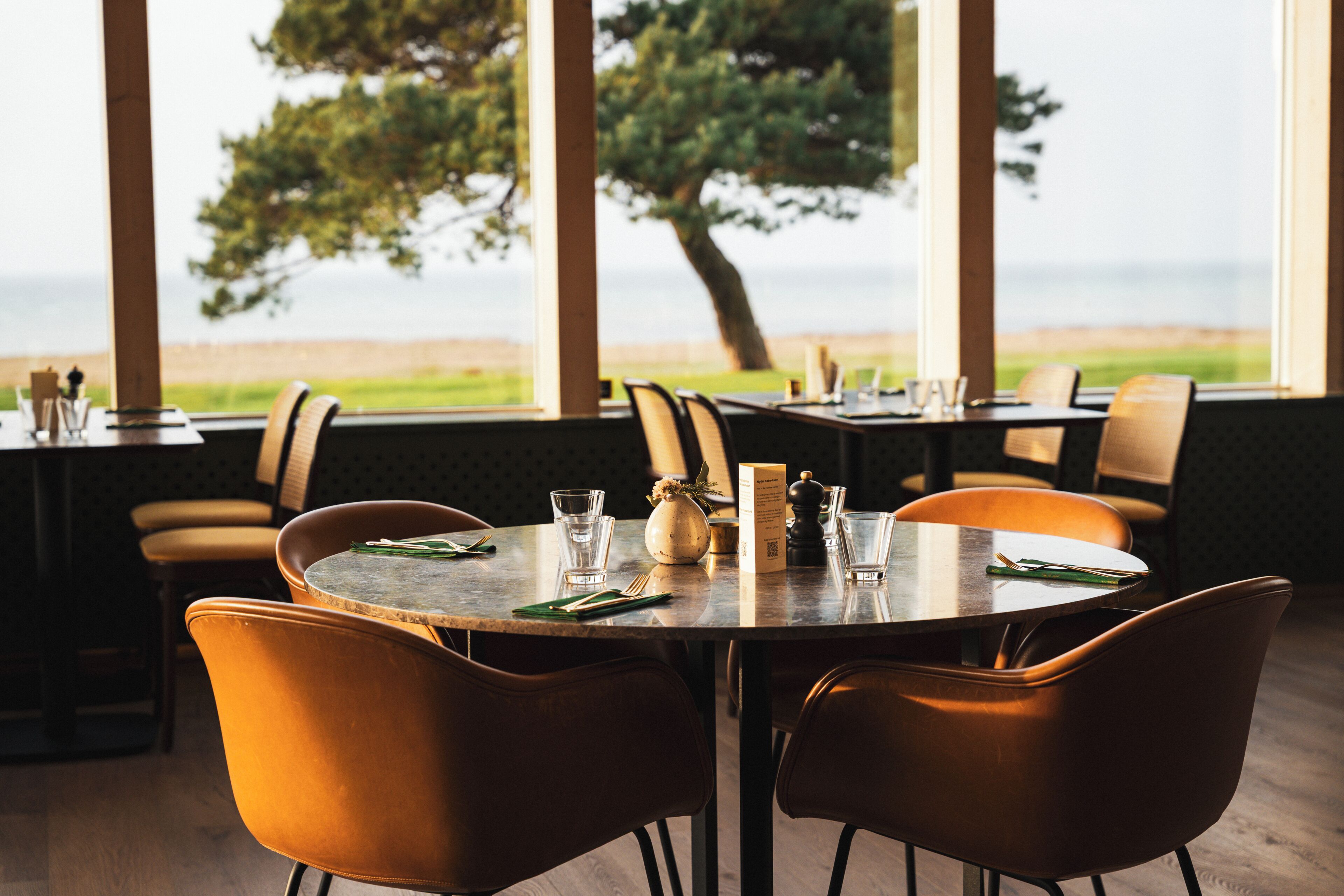A restaurant with a view of the sea, featuring round tables, brown leather chairs, and a simple table setting with a menu.