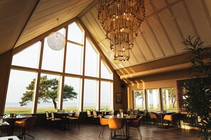 A restaurant with large windows, wooden tables, and chairs, and a hanging light fixture. - Barsebäck Resort Hotell (Loddekopinge)