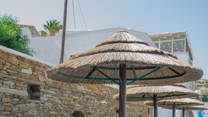 A row of wooden lounge chairs with white cushions and yellow towels, arranged under thatched umbrellas, against a stone wall with a clear blue sky above. - Golden Sun Hotel (Ios)