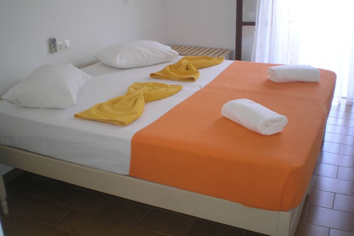 A neatly made bed with white and orange linens, a yellow towel, and a white towel.