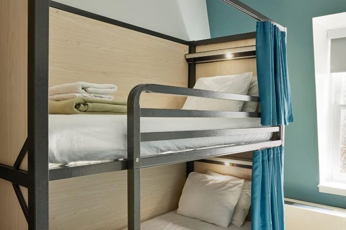 Een stapelbed met een houten hoofdeinde, een raam met gordijnen en uitzicht op een gebouw.