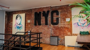 Una habitación con pared de ladrillo, un cartel grande que dice 'NYC', una escalera y una mesa.