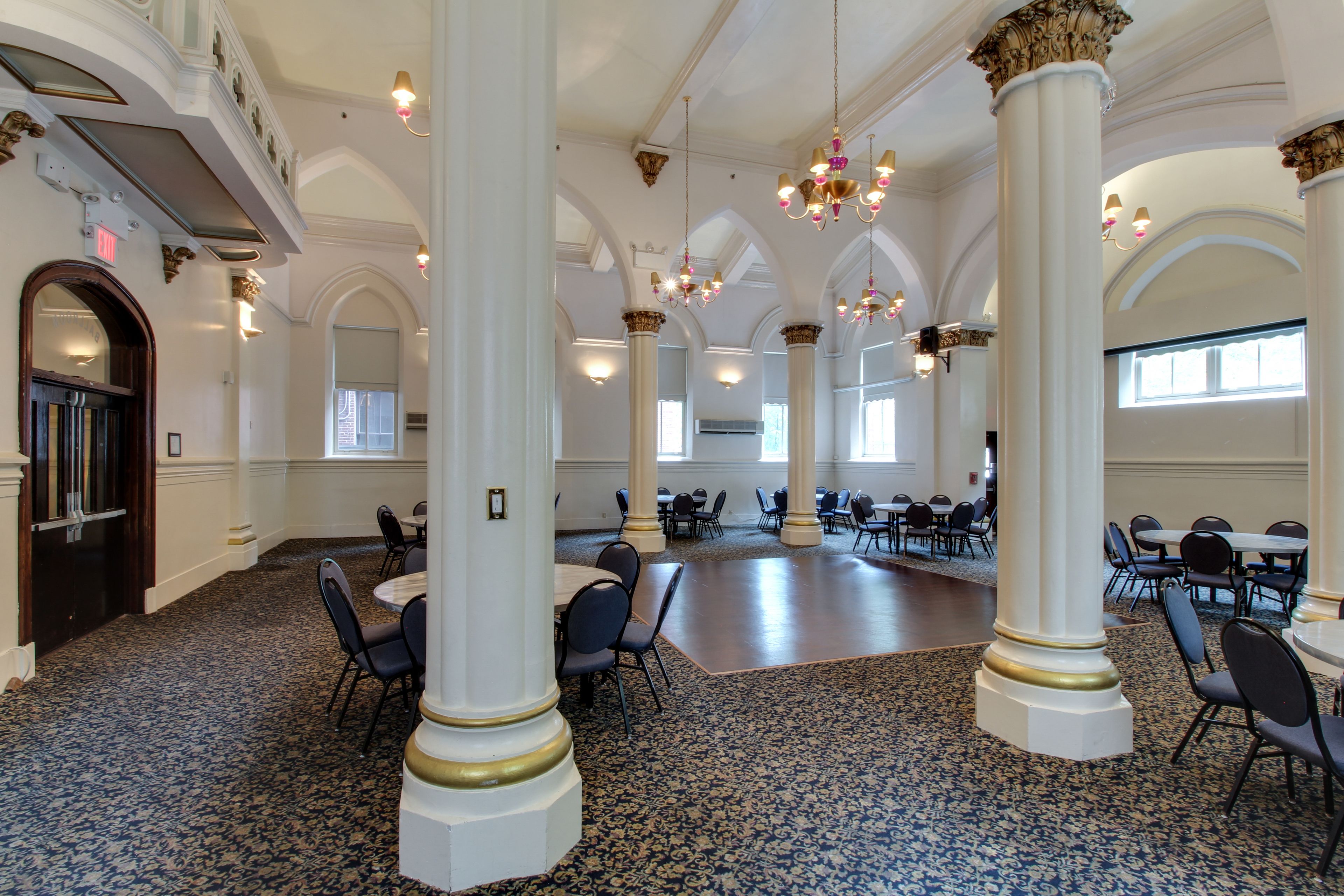 Une vaste salle dotée de hauts plafonds, de colonnes richement décorées et de lustres. Des tables et des chaises y sont disposées pour accueillir des événements.