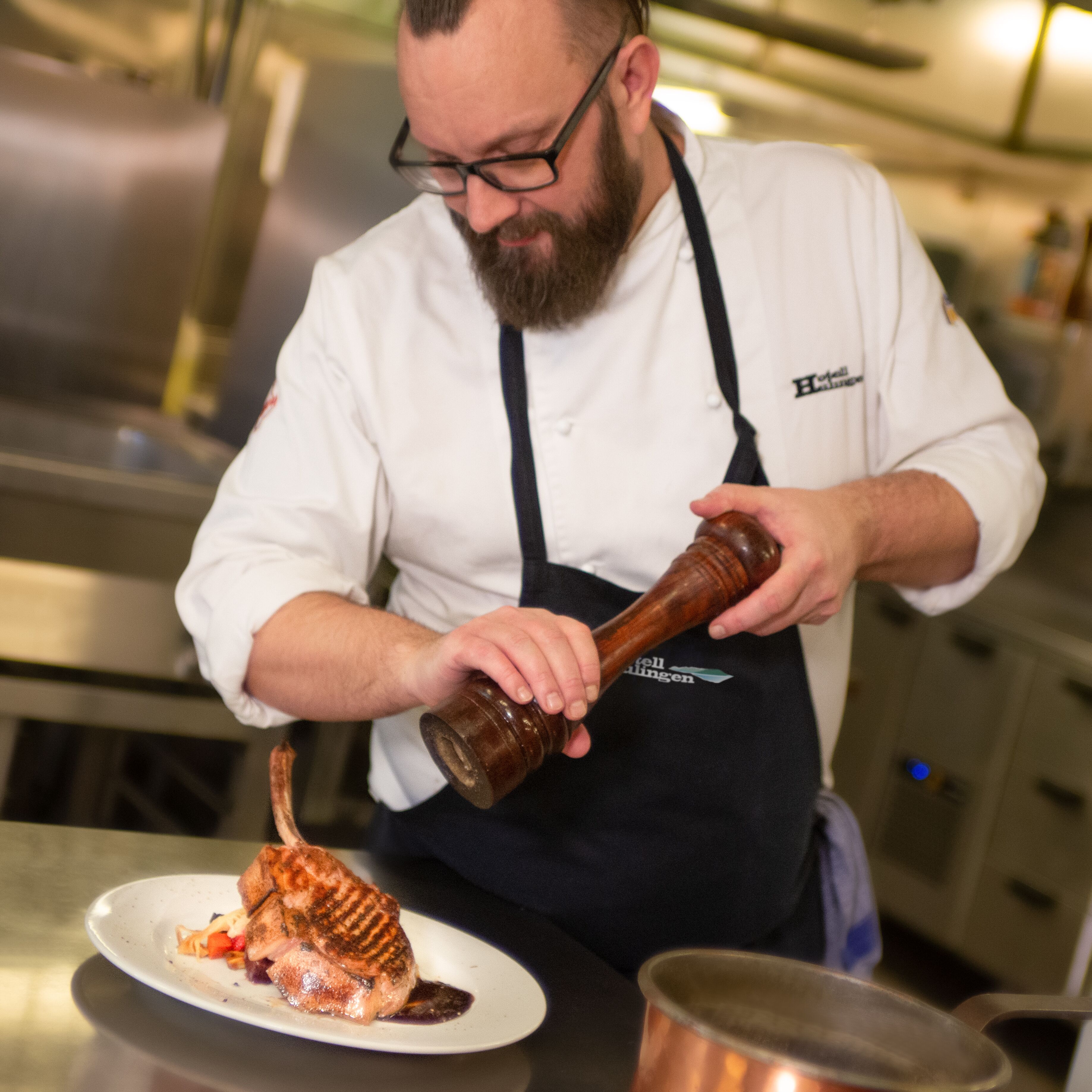 Un chef con uniforme blanco y delantal negro sirve salsa de una botella sobre un plato de comida.