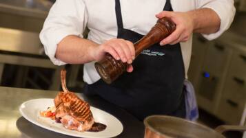 Un chef con uniforme blanco y delantal negro sirve salsa de una botella sobre un plato de comida.