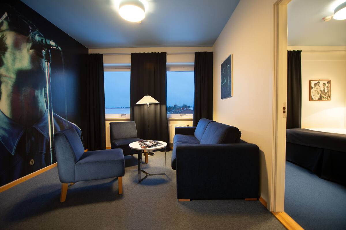 A modern hotel room with a large window, a sofa, a chair, a small table, and a lamp.