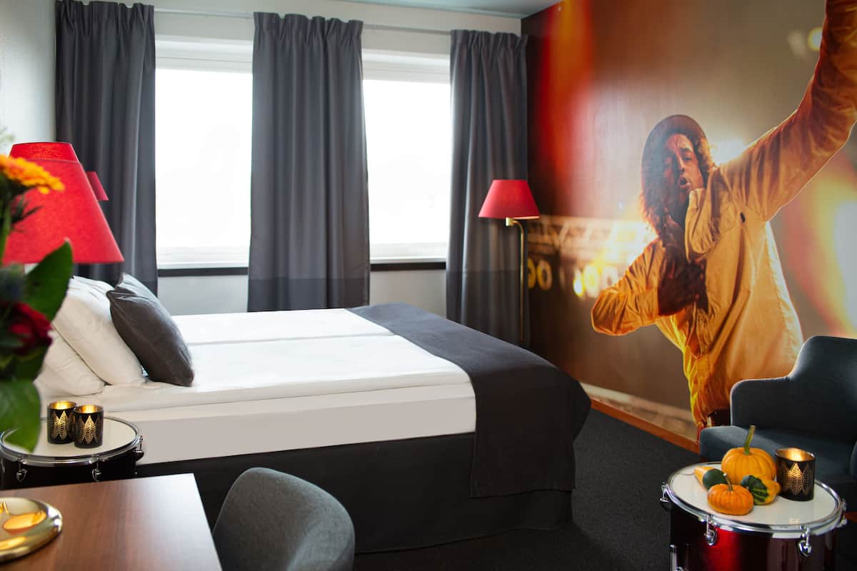 A hotel room with a bed, a desk with a lamp, a chair, and a wall with a poster.