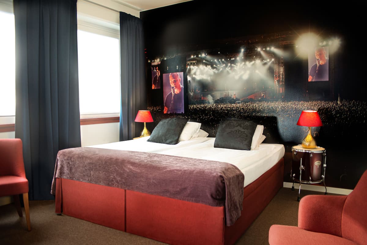 A hotel room with a large bed, two red chairs, a desk, and a large screen displaying a concert.