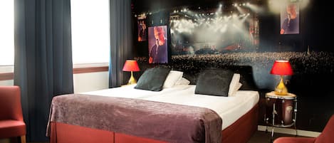 A hotel room with a large bed, two red chairs, a desk, and a large screen displaying a concert.
