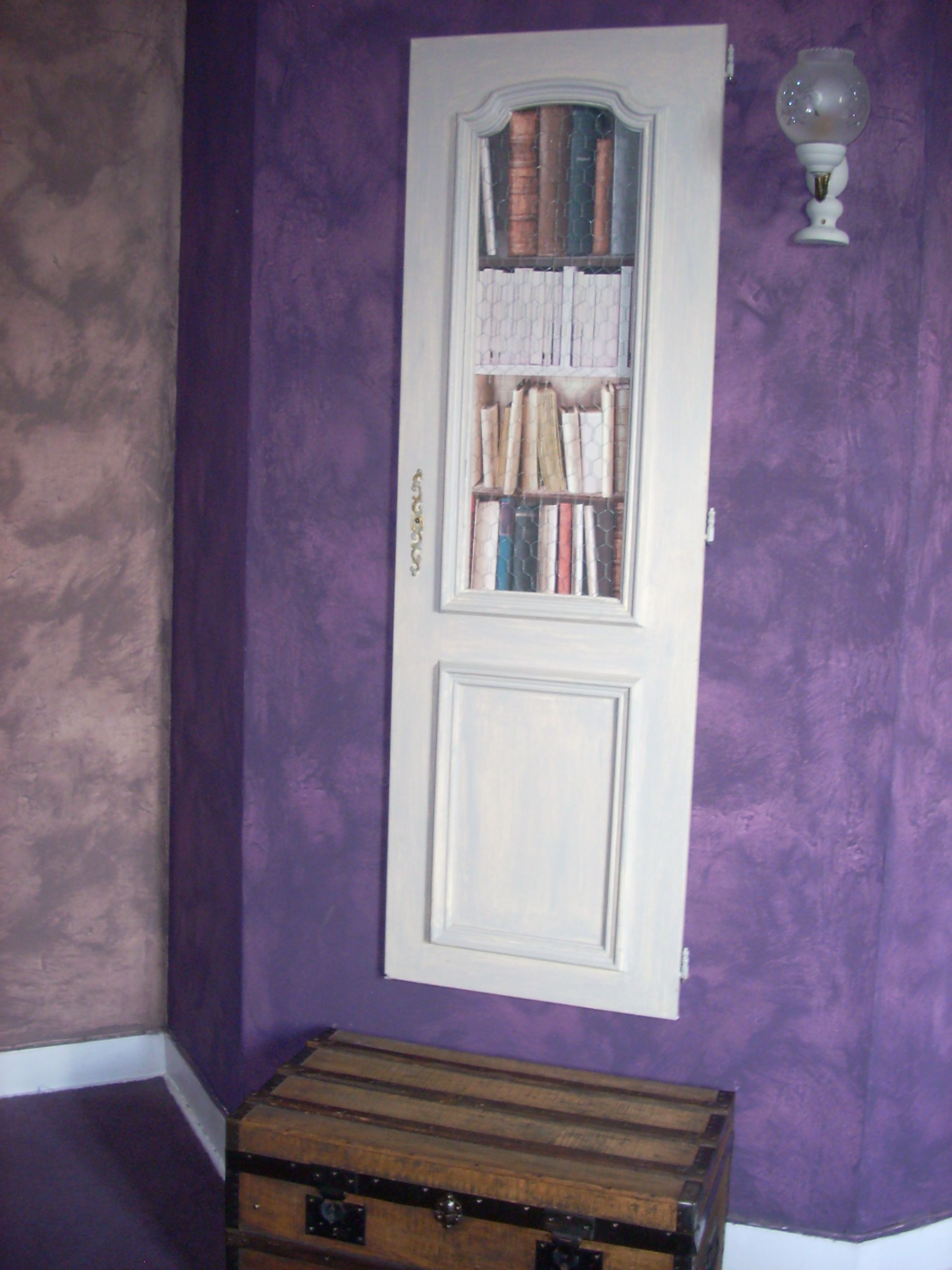 Ein Zimmer mit violetten Wänden, einer weißen Tür mit einem Bücherregal und einer Holzkiste.