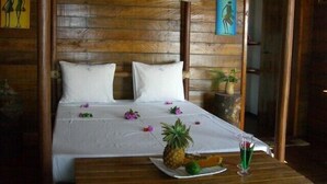 A wooden room with a bed, a table with a pineapple and a drink, and a door. - Riake Resort & Villa (Nosy Boraha)
