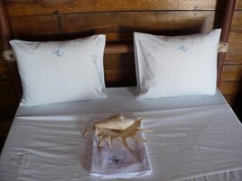 Cama con sábanas blancas y dos almohadas, cabecera de madera y una concha decorativa sobre la colcha.