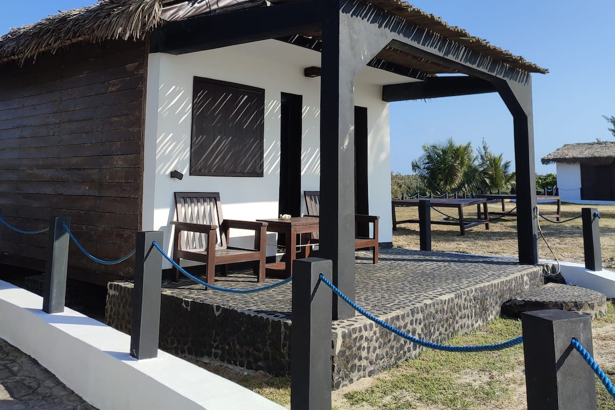 A small, rustic building with a thatched roof and a covered patio area. It features a stone wall and a wooden fence with a blue rope barrier.