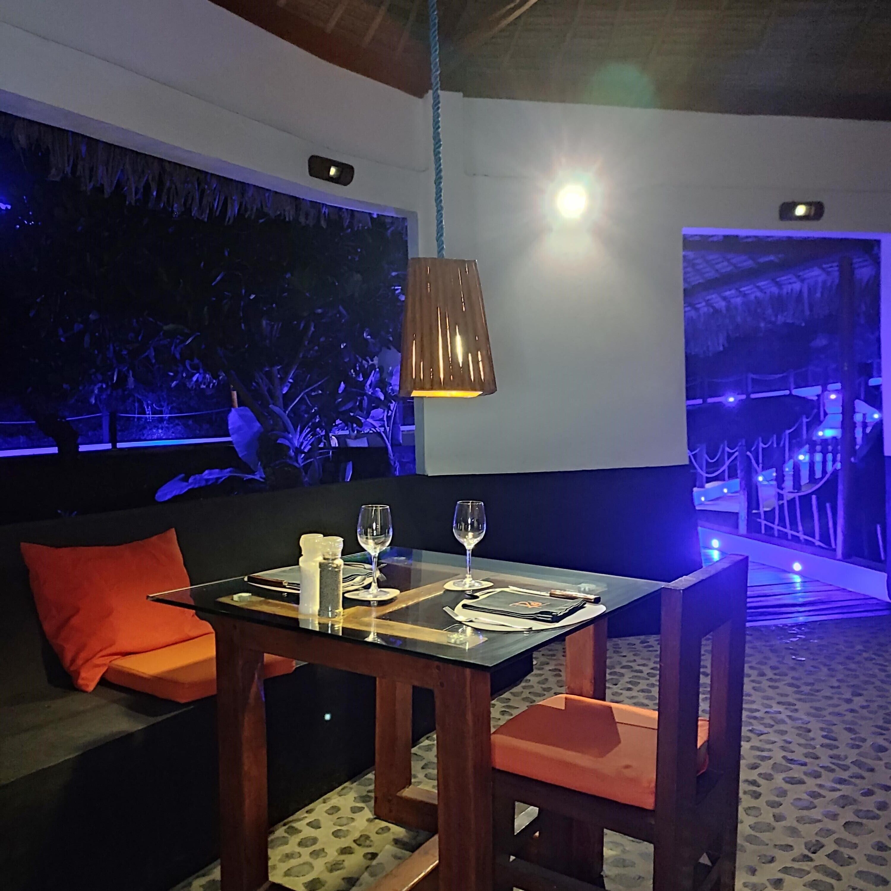 Un coin repas avec une table dressée pour deux personnes, un canapé avec des coussins orange et un luminaire suspendu.