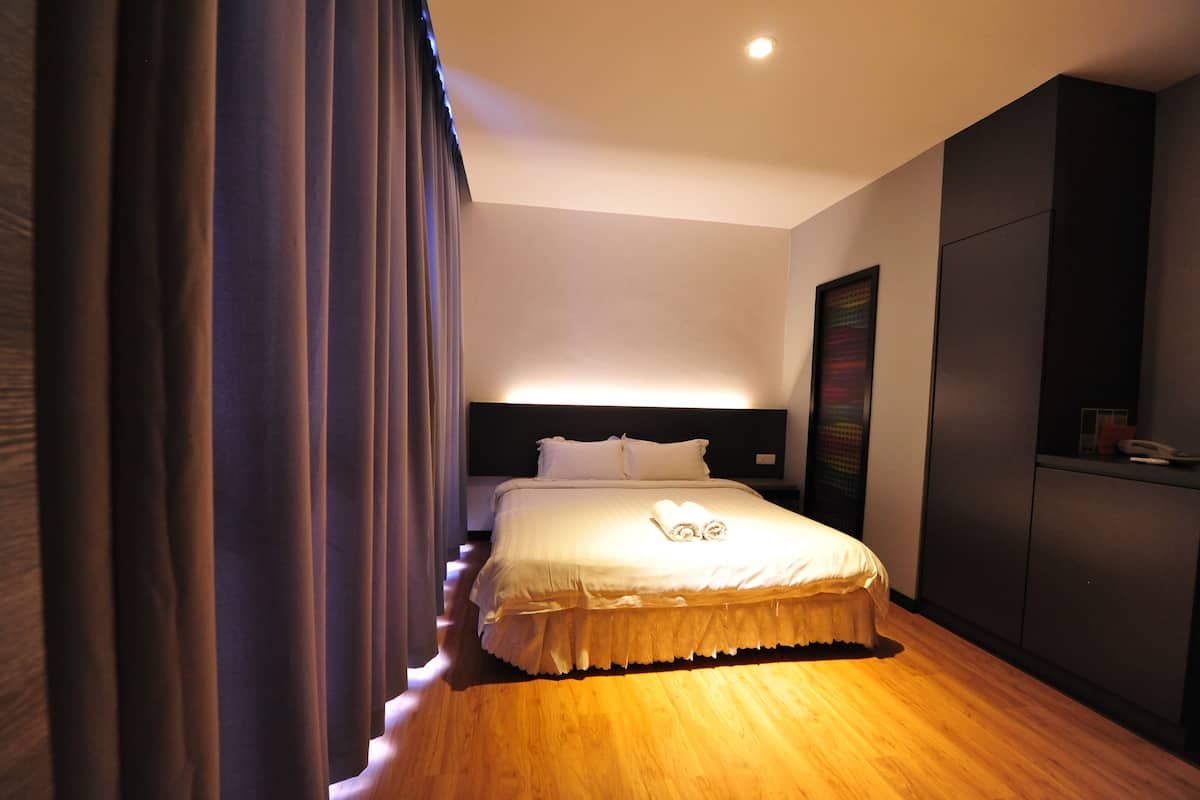 Una cama bien hecha con una colcha blanca y almohadas, suelo de madera y cabecero oscuro.