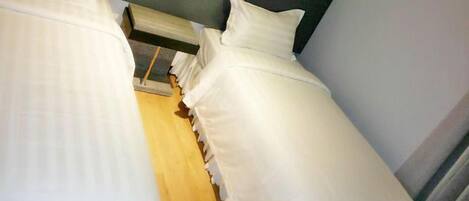 Habitación de hotel estrecha con dos camas individuales, piso de madera y cabecera oscura.